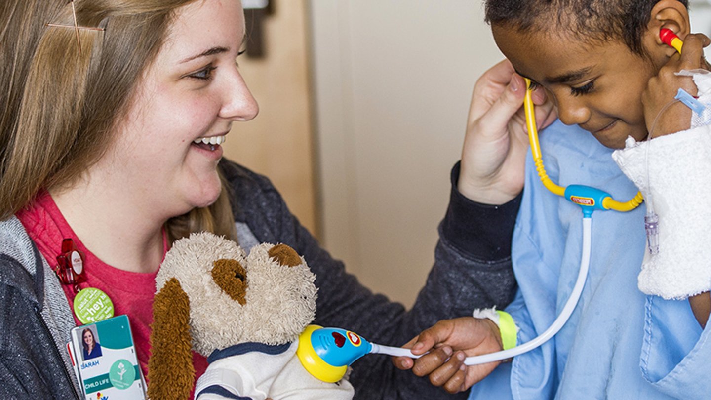 A child life specialists helps a kid listen to a stuffed animal's heartbeat with a toy stethoscope.