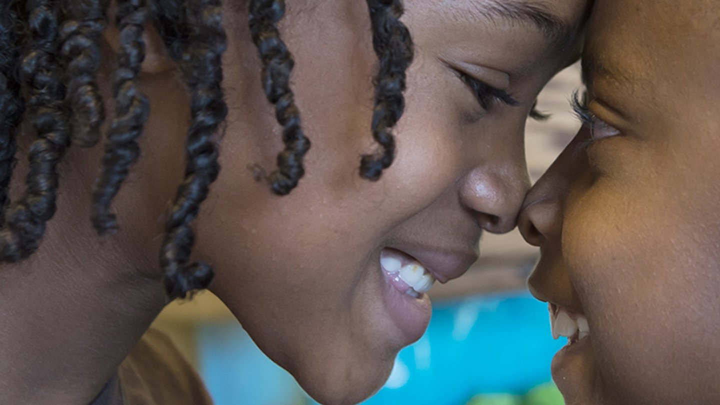 A close-up of two kids smiling and touching their foreheads and noses to each other.