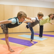 A woman wearing a yellow blouse and black pants stands barefoot in airplane pose on a light blue yoga mat. To her right are two boys wearing collared shirts under a vest and copying her same pose on purple and pink yoga mats. Both boys are barefoot. One is wearing jeans and the other is wearing plaid shorts.