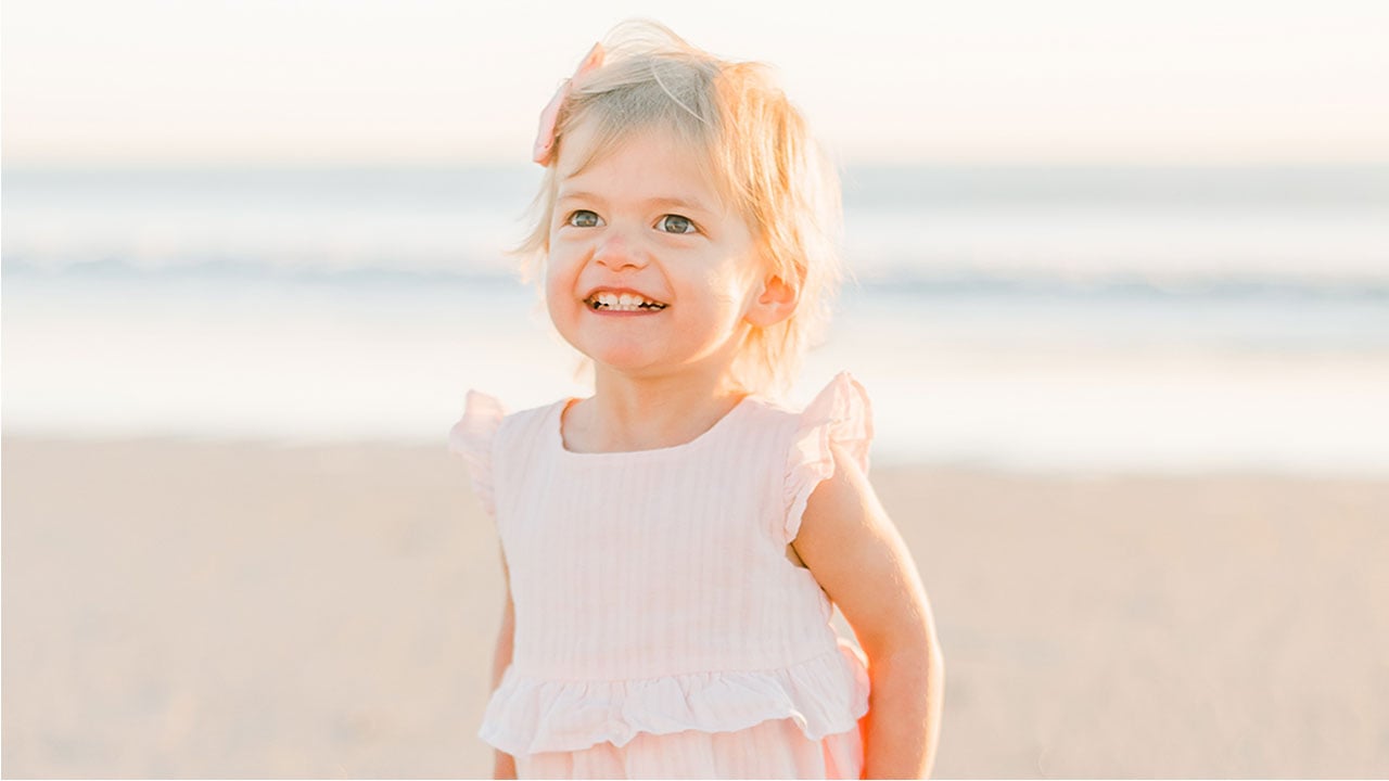 A toddler girl with blonde hair stands on the beach, smiling. She received care for fetal heart block at Children’s Colorado.