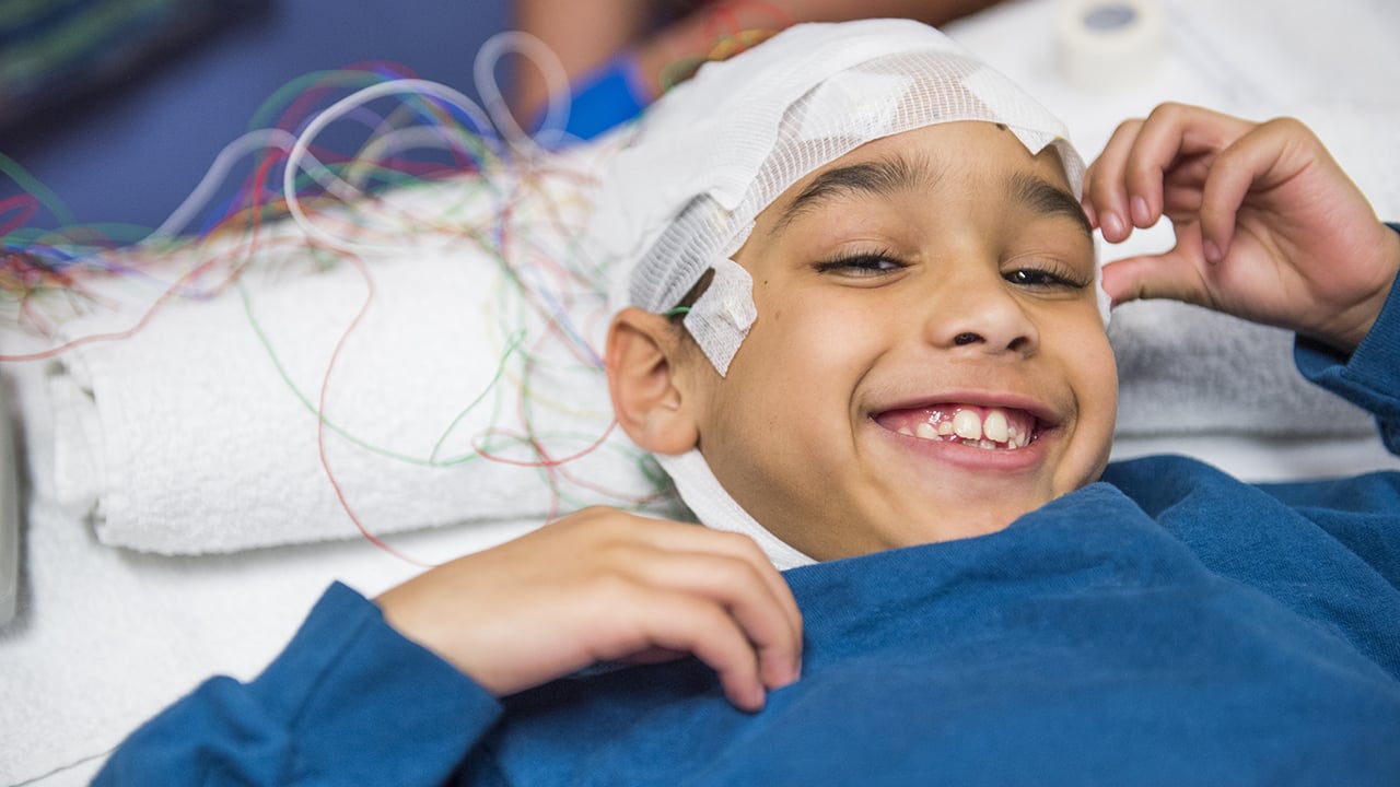 A boy in a blue shirt lies in a bead with his head wrapped in bandages and hooked up to sensors