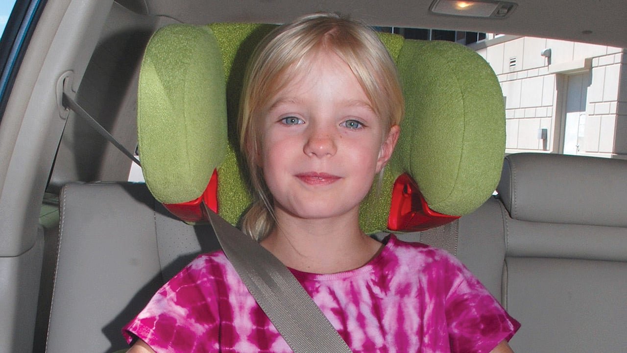 A girl with blonde hair in a ponytail sits in a lime green booster seat with a seatbelt across her shoulder inside a car with gray interior