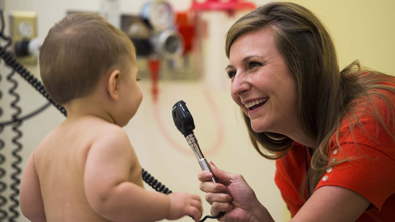 A woman with long brown hair and wearing a red shirt holds up a light instrument in front of a baby with short brown hair.
