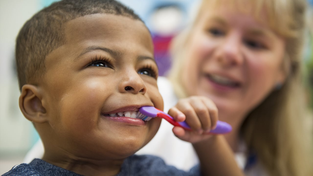 A young boy wearing a blue shirt smiles and brushes his teeth with a purple and pink toothbrush while a doctor with long blonde hair watches in the background.