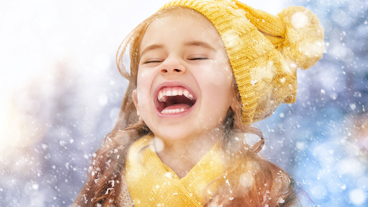 A close-up of a girl with long brown hair, wearing a yellow knit hat and a yellow scarf, laughing as snow falls down around her.