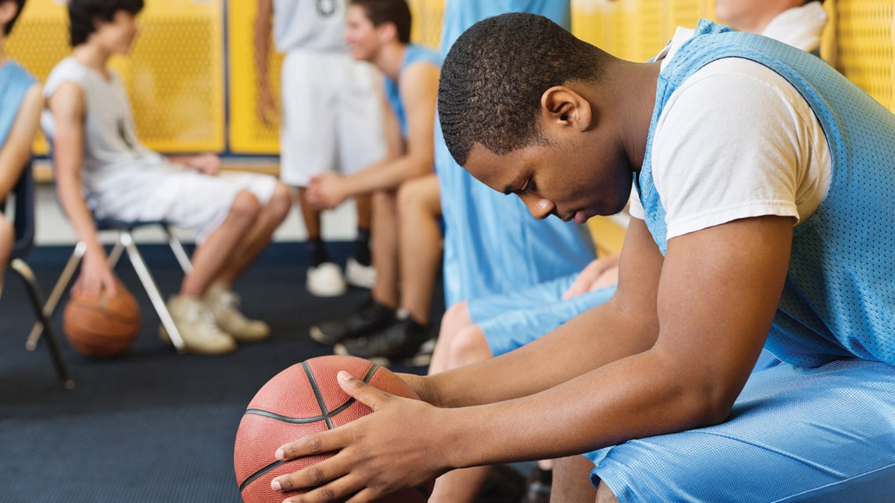 A basketball player sits on a bench in the locker room holding a basketball and looking down while his teammates look happy.