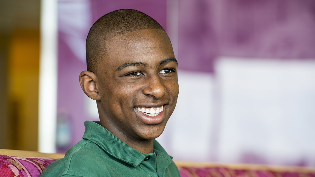 A teenage boy wearing a green polo sits on a magenta couch smiling.