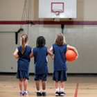 Three girls in navy blue jerseys stand on a basketball court holding a ball and looking at the basket.